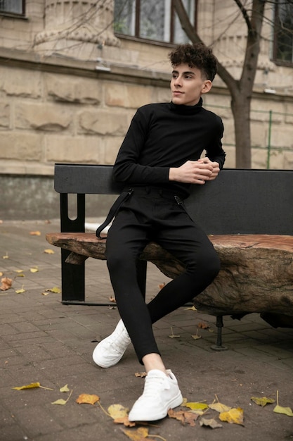 Junger Mann mit moderner Frisur in schwarz gekleidet sitzt entspannt auf einer Bank und schaut zur Seite