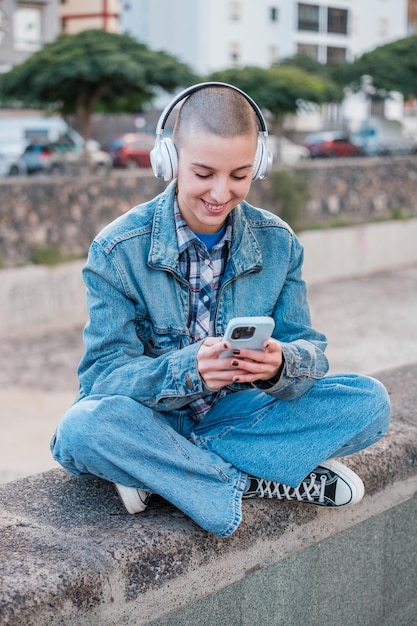 Junger Mann mit rasierten Haaren, der mit seinem Smartphone interagiert, während er Musik hört, sitzt in der Stadt Concept Lifestyle Technology Slide