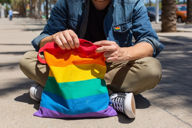 Junger Mann mit Regenbogen-Mehrwegbeutel, der auf Bürgersteig sitzt