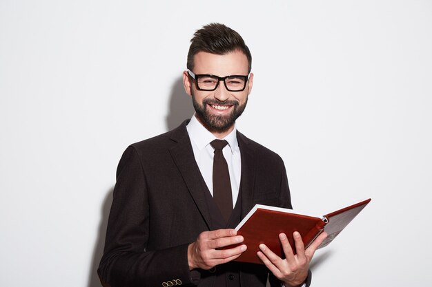 Junger Mann mit schwarzen Haaren und Bart, die weißes Hemd, Anzug und Sonnenbrille am weißen Studiohintergrund tragen, glücklich, ein Buch lesend.