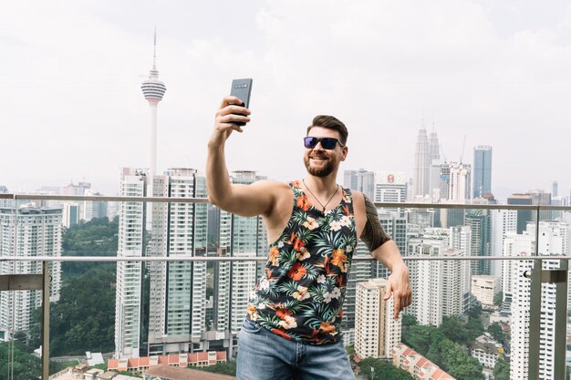 Junger Mann mit Sonnenbrille und einem ärmellosen T-Shirt mit Blumendruck, das ein Selfie mit der Stadt macht