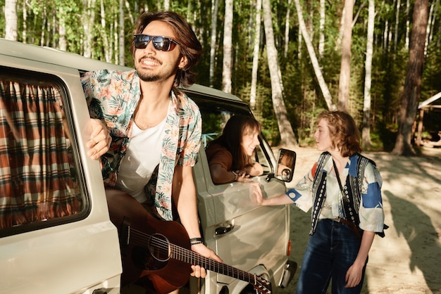 Junger Mann mit Sonnenbrille und Gitarre, der mit seinen Freunden im Wald reist