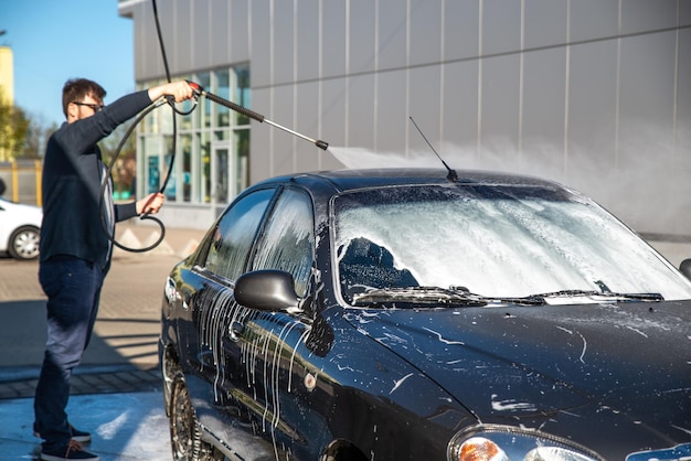 Junger Mann mit Sonnenbrille wäscht Auto in der Selbstbedienungswaschanlage