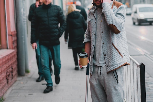 Junger Mann mit Stock in lässiger Kleidung steht auf der Straße der Stadt Stock Menschenmenge Hauptverkehrszeit warten Tasse halten Kaffee Leben Bein Junge Schritt zu Fuß Kerl Krankheit Mann allein urban