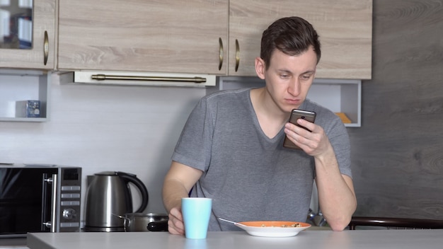 Junger Mann mit Telefon isst an einem Tisch in der Küche.