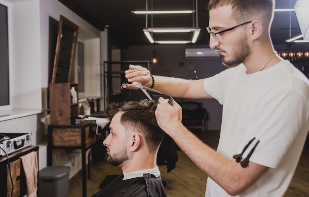 Junger Mann mit trendigem Haarschnitt am Friseurladen. Barber macht die Frisur und Bart schneiden.