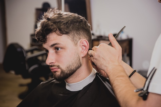 Junger Mann mit trendigem Haarschnitt am Friseurladen. Barber macht die Frisur und Bart schneiden.