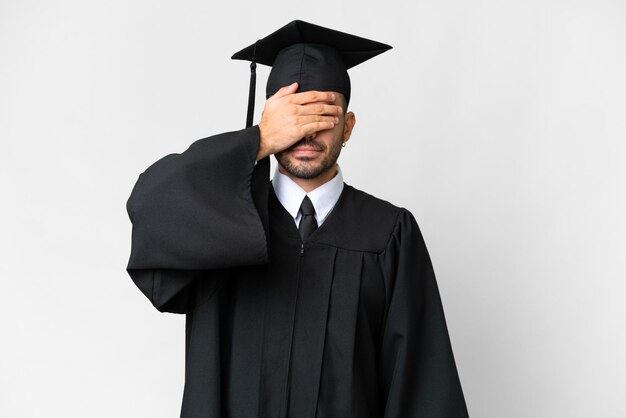 Junger Mann mit Universitätsabschluss über isoliertem weißem Hintergrund, der die Augen mit den Händen bedeckt. Will nichts sehen