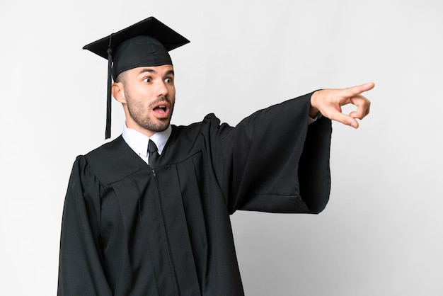 Junger Mann mit Universitätsabschluss über isoliertem weißem Hintergrund, der weg zeigt