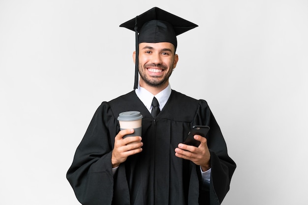 Junger Mann mit Universitätsabschluss vor isoliertem weißem Hintergrund mit Kaffee zum Mitnehmen und einem Handy