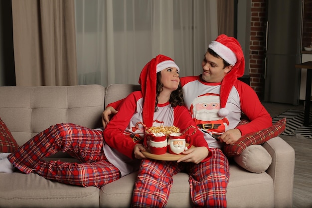 junger Mann mit Weihnachtsmannmütze sitzt auf der Couch und umarmt seine Freundin im Weihnachtspyjama