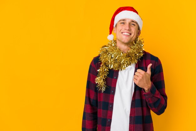 Junger Mann mit Weihnachtsmütze hält ein Geschenk lokalisiert auf gelber Wand lächelnd und hebt Daumen hoch