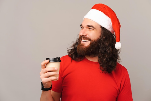 Junger Mann mit Weihnachtsmütze hält eine Tasse Kaffee zum Mitnehmen und schaut zur Seite