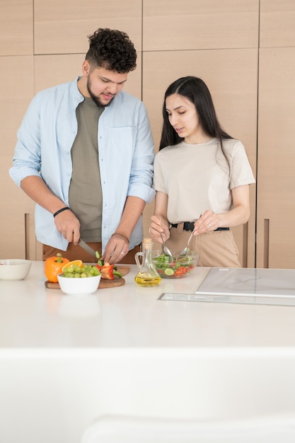 Junger Mann schneidet Gemüse für Salat, während seine Frau Zutaten in einer Schüssel mischt