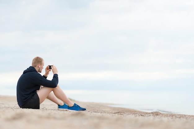 Junger Mann sitzt am Strand nach dem Joggen und fotografiert das Meer und schaut auf das Telefon