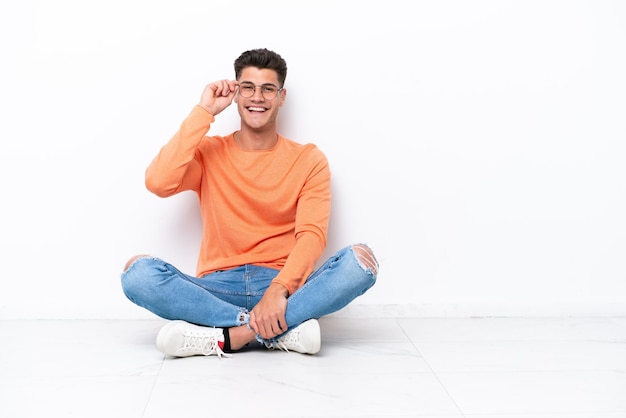 Junger Mann sitzt auf dem Boden isoliert auf weißem Hintergrund mit Brille und glücklich