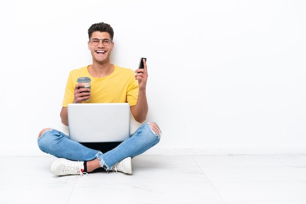 Junger Mann sitzt auf dem Boden isoliert auf weißem Hintergrund und hält Kaffee zum Mitnehmen und ein Handy