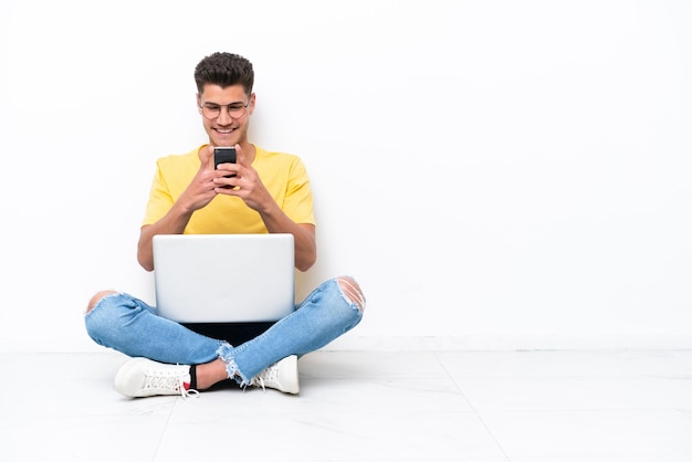 Junger Mann sitzt auf dem Boden isoliert auf weißem Hintergrund und sendet eine Nachricht mit dem Handy
