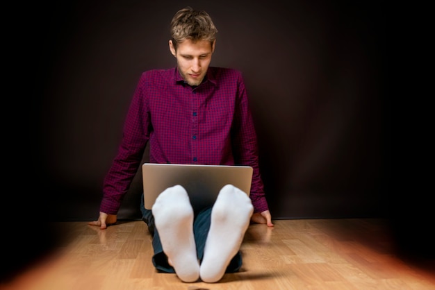 Junger Mann sitzt auf dem Boden und benutzt Laptop auf dunklem Hintergrund b