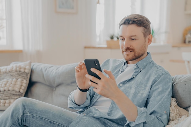 Junger Mann sitzt auf der Couch und surft mit seinem Handy im Internet