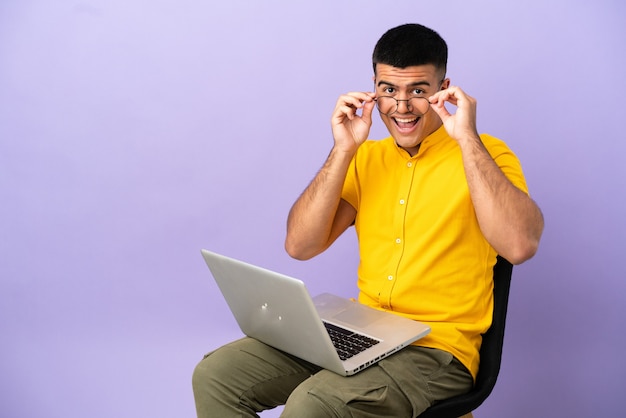 Junger Mann sitzt auf einem Stuhl mit Laptop mit Brille und überrascht