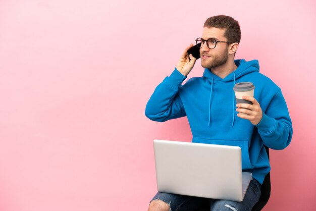 Junger Mann sitzt auf einem Stuhl mit Laptop mit Kaffee zum Mitnehmen und einem Handy