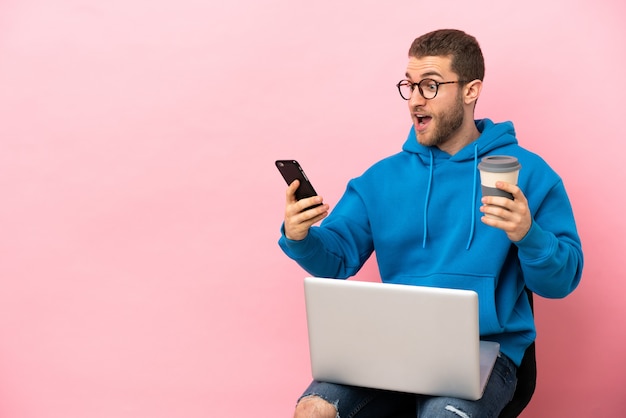 Junger Mann sitzt auf einem Stuhl mit Laptop mit Kaffee zum Mitnehmen und einem Handy