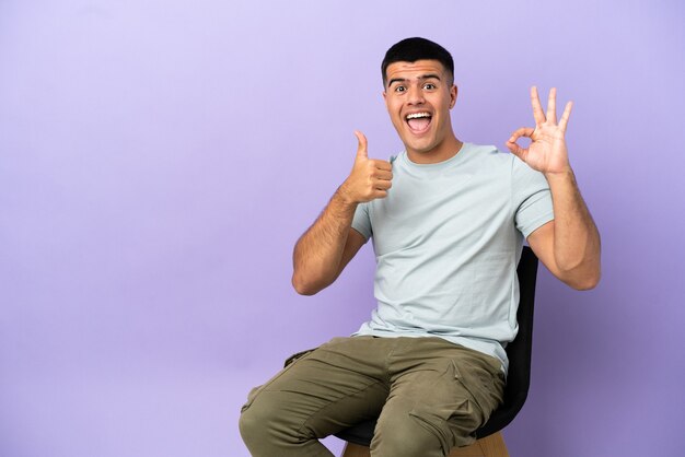 Junger Mann sitzt auf einem Stuhl über isoliertem Hintergrund und zeigt ein OK-Zeichen und eine Daumen-Hoch-Geste