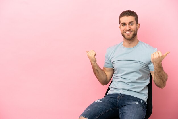 Junger Mann sitzt auf einem Stuhl über isoliertem rosa Hintergrund mit Daumen hoch Geste und lächelt