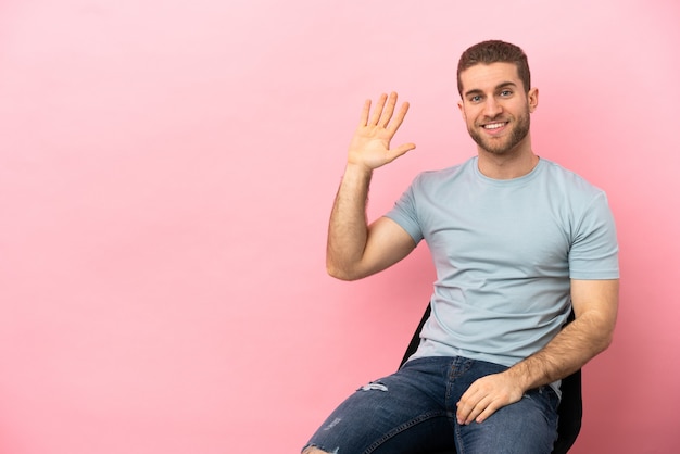 Junger Mann sitzt auf einem Stuhl über isoliertem rosa Hintergrund und grüßt mit der Hand mit glücklichem Ausdruck