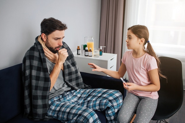 Junger Mann sitzt auf Sofa und hustet. Sein Hals war mit einem Schal bedeckt. Die Decke liegt auf den Schultern. Kleines Mädchen macht sich Sorgen um ihn. Sie hält das Thermometer in der Hand.