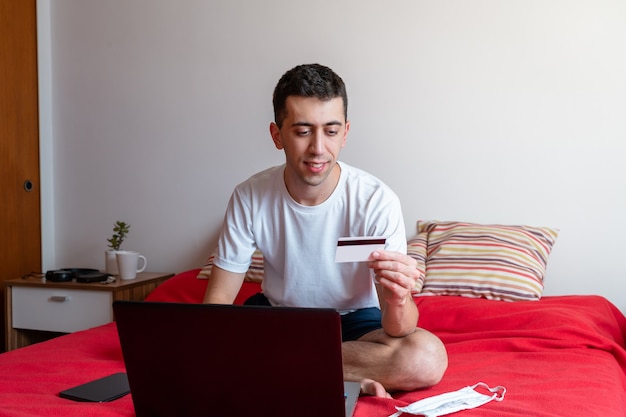 Junger Mann sitzt im Bett beim Online-Shopping und zahlt mit seiner Kreditkarte