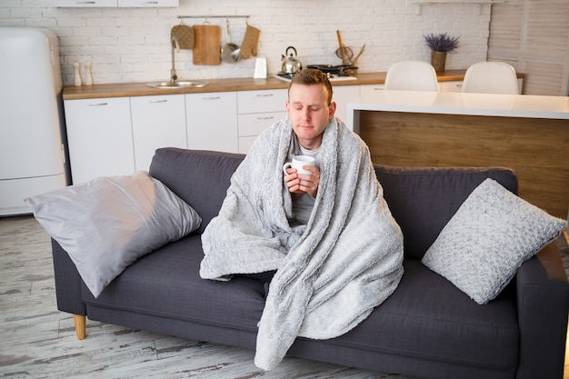 Junger Mann sitzt mit einer in eine Decke gewickelten Tasse Tee auf dem Sofa und ist krank