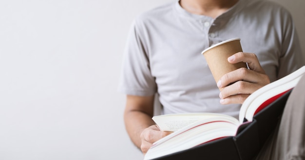 Junger Mann sitzt und liest ein Buch, während er eine Pappbecher Kaffee im Raum hält