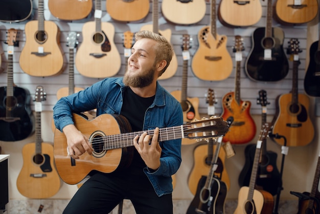 Junger Mann spielt auf akustischer Gitarre im Musikgeschäft. Sortiment im Musikinstrumentengeschäft, männlicher Musiker kauft Ausrüstung