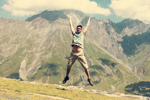 Junger Mann springt mit erhobenen Händen gegen den Himmel und die Berge