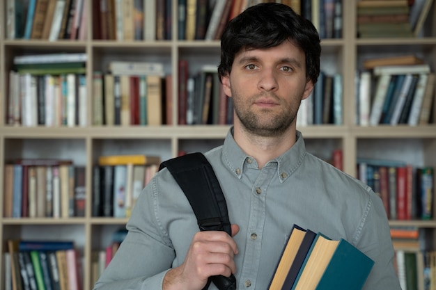 Junger Mann steht mit Büchern und einem Rucksack in der Bibliothek