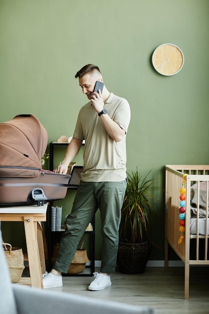 Junger Mann telefoniert mit dem Handy, während er sich zu Hause um sein neugeborenes Baby in der Wiege kümmert