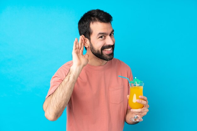 Junger Mann über das Halten eines Cocktails über isolierte blaue Wand, die etwas hört, indem Hand auf das Ohr legt