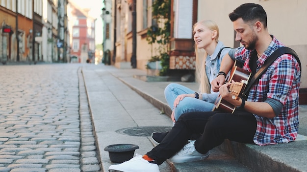 Junger Mann und Frau der Straßensänger, die auf Bürgersteig sitzen, Gitarre spielen und singen. Lifestyle-Konzept.