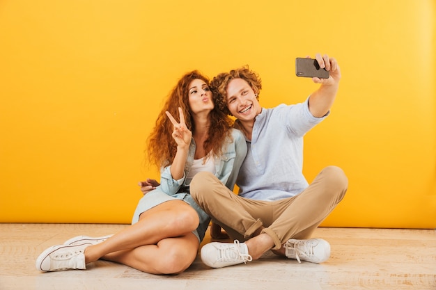 Junger Mann und Frau des optimistischen Paares, die auf Boden zusammen sitzen und Friedenszeichen zeigen, während sie selfie auf Smartphone nehmen, lokalisiert über gelbem Hintergrund