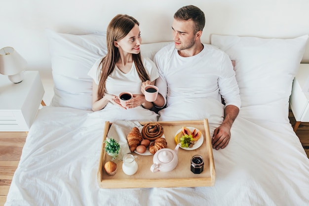 Junger Mann und Frau, die Frühstück in einem weißen Bett im Schlafzimmer haben