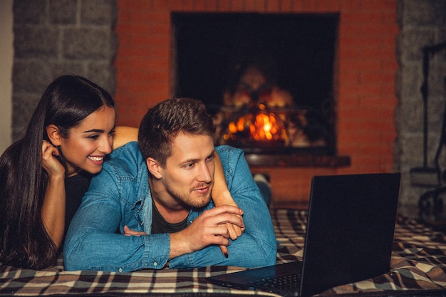 Junger Mann und Frau liegen zusammen auf einer Decke auf dem Boden. Sie sehen sich Filme auf dem Laptop an. Paar ist glücklich. Sie sind in der Nähe des Kamins