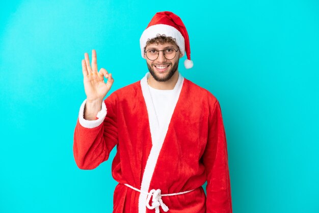 Junger Mann verkleidet als Weihnachtsmann auf blauem Hintergrund isoliert, der ein OK-Zeichen mit den Fingern zeigt