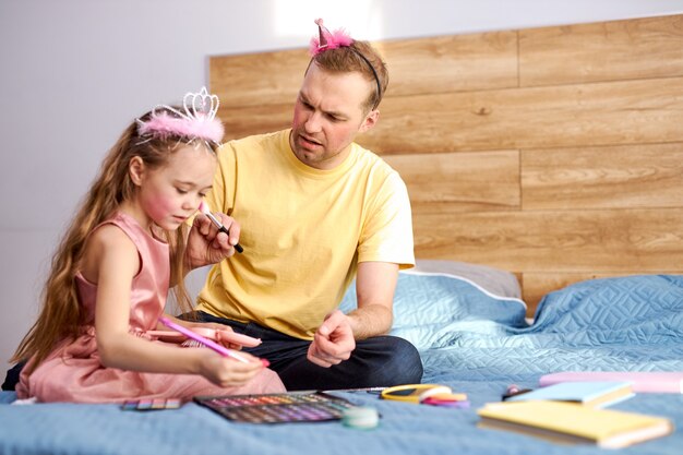 junger Mann zu Hause mit seinem kleinen süßen Mädchen, Make-up auf Gesicht mit dekorativen Mutterkosmetik.