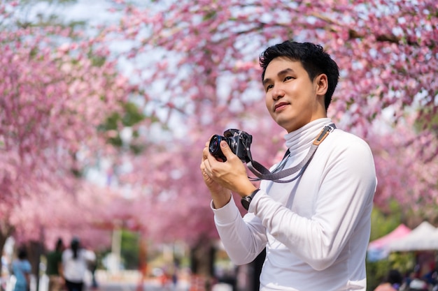 Junger Mannreisender, der Kirschblüten oder Sakura-Blume schaut, die blüht und Kamera hält, um ein Foto im Park zu machen