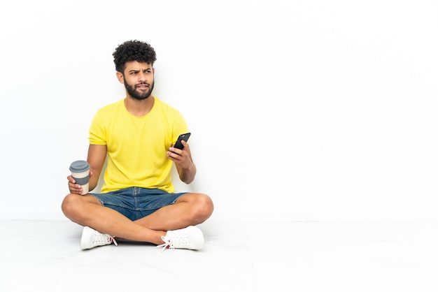 Junger marokkanischer hübscher Mann, der auf dem Boden über lokalisiertem Hintergrund hält, der Kaffee hält, um und ein Handy wegzunehmen, während er etwas denkt