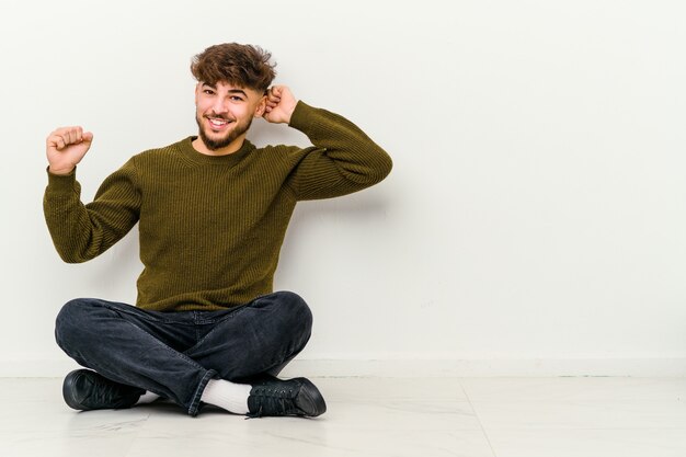 Junger marokkanischer Mann, der auf dem Boden lokalisiert auf weißem Wandtanzen sitzt und Spaß hat.
