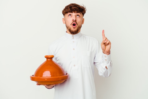 Junger marokkanischer Mann, der das typische arabische Kostüm hält, das eine Tajine hält, die oben mit geöffnetem Mund zeigt.