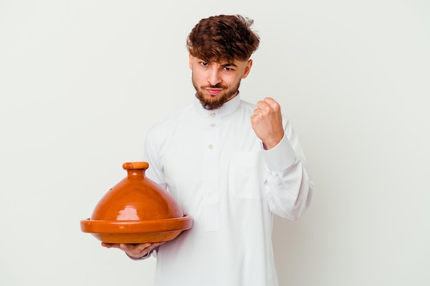 Junger marokkanischer Mann, der das typische arabische Kostüm hält, das eine Tajine lokalisiert auf weiß zeigt Faust zur Kamera, aggressiven Gesichtsausdruck zeigt.